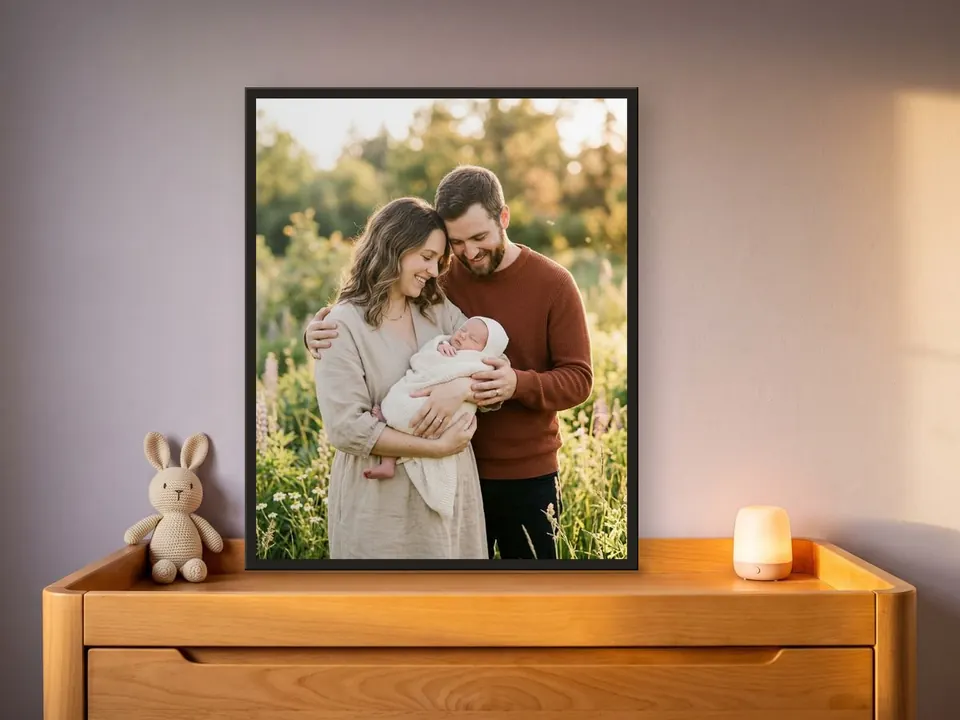Birth print in golden light, a couple with their newborn in a black frame, on a beech changing chest against a dusty lilac wall, with a knitted bunny and a night light